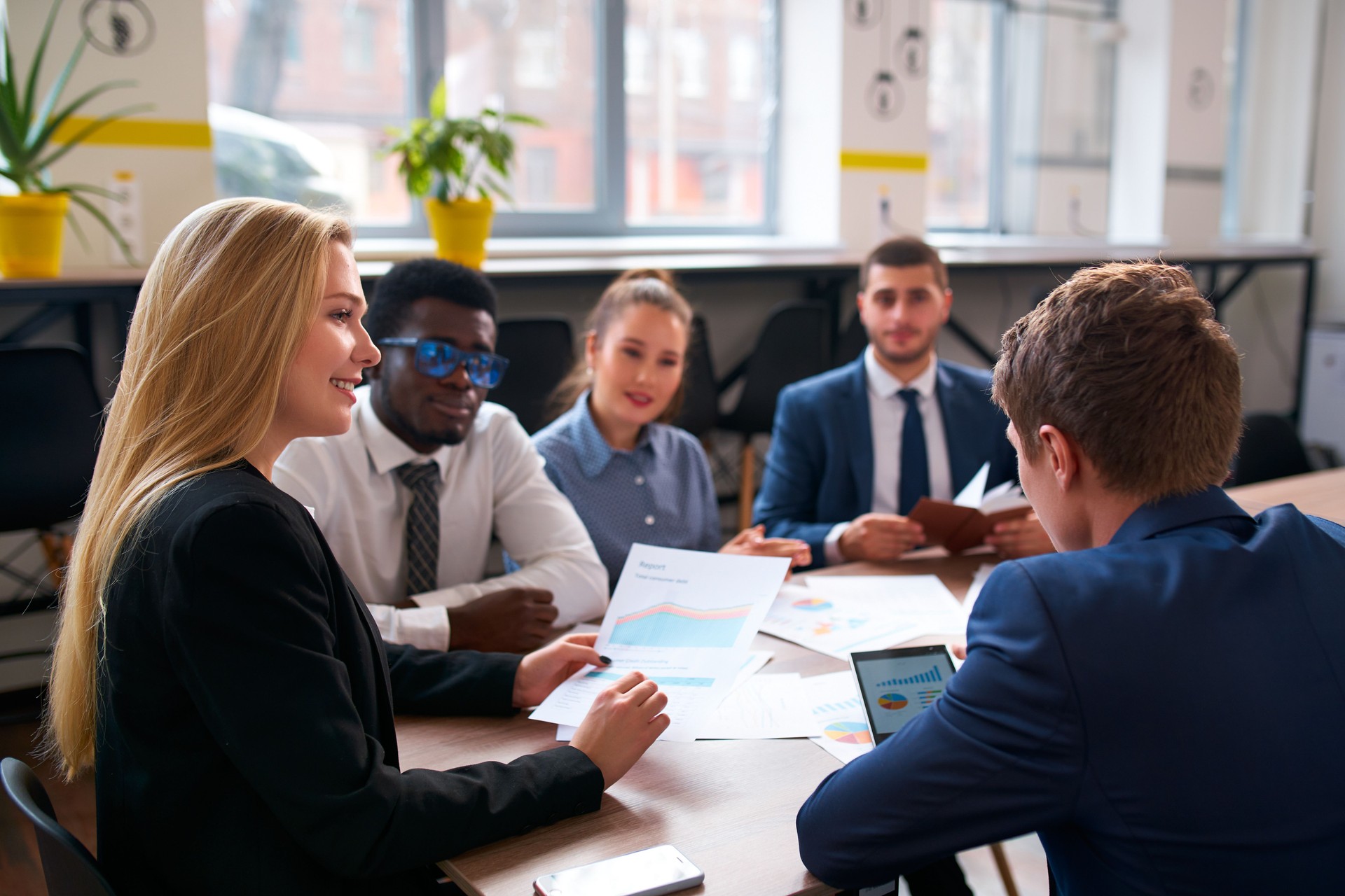 Multiracial team in office analyzes charts for project. Diverse group includes Asian boss, African businessman, discussing strategy. Multinational employees involved in planning, using diagrams.